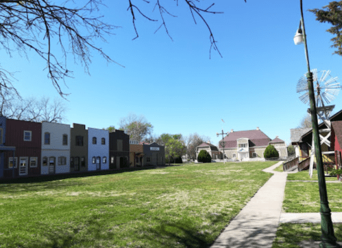 A grassy area with a pathway, surrounded by small buildings and a windmill under a clear blue sky.
