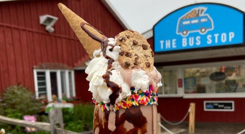 A decadent ice cream cone topped with whipped cream, chocolate syrup, sprinkles, and a cookie, in front of a red building.