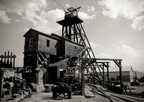 Black and white image of an old mining structure with a tall headframe and wooden buildings against a cloudy sky.