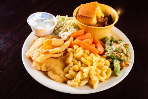 A plate of food featuring fried fish, macaroni, green beans, carrots, coleslaw, cornbread, and a side of sauce.