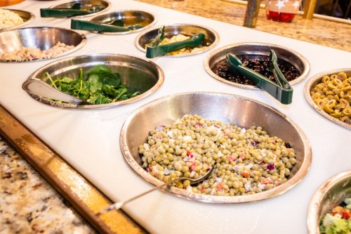 A variety of salad ingredients in metal bowls on a countertop, including greens, beans, and a mixed grain salad.