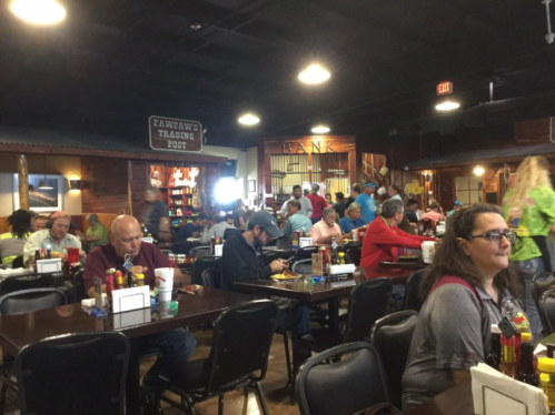 A busy restaurant interior with patrons dining at tables, colorful decor, and a bank-themed area in the background.