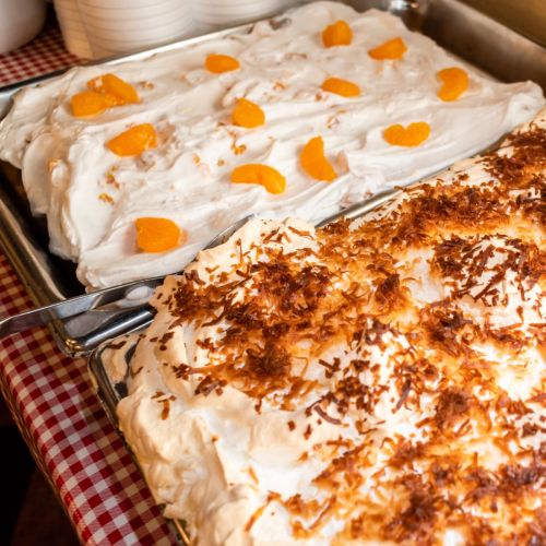 Two trays of dessert: one topped with orange slices and the other with toasted coconut, on a checkered tablecloth.