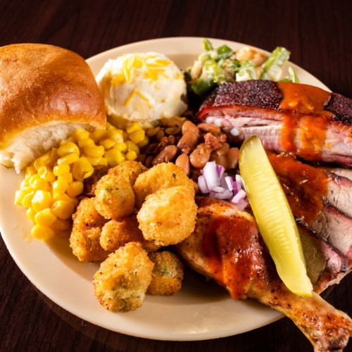 A plate of barbecue featuring brisket, chicken, corn, okra, beans, mashed potatoes, and a roll, garnished with pickles.