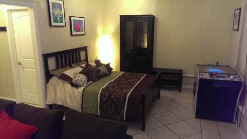 A cozy bedroom featuring a bed with decorative pillows, a dresser, and a small table, with soft lighting and tiled floor.