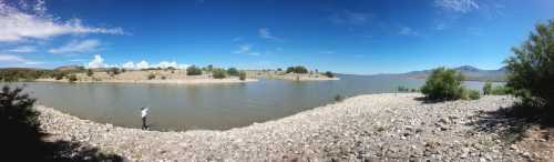 A panoramic view of a calm lake surrounded by rocky shores and green vegetation under a clear blue sky.