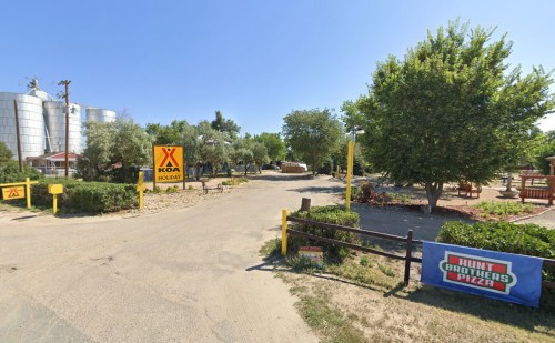 A KOA campground entrance with trees, a dirt road, and a sign for Hunt Brothers Pizza in a sunny outdoor setting.