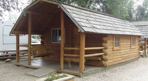 A rustic log cabin with a porch and swing, surrounded by gravel and trees.