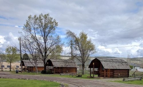Three rustic wooden cabins surrounded by trees and a gravel path under a cloudy sky.