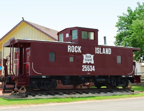 A red caboose labeled "Rock Island 25534" parked beside a building, surrounded by greenery.