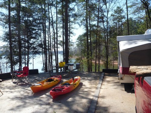 A serene campsite by a lake, featuring two kayaks, a picnic table, and surrounded by tall trees.