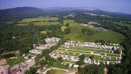 Aerial view of a campground with rows of RVs, surrounded by green fields and distant mountains under a clear blue sky.