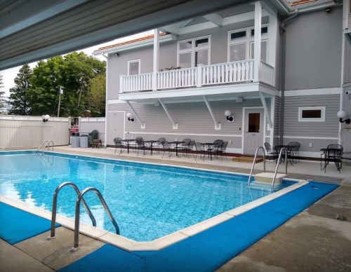 A clear swimming pool surrounded by a patio and chairs, with a two-story gray building in the background.