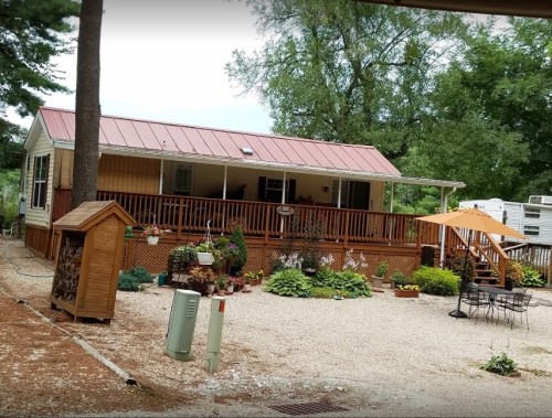 A cozy mobile home with a red metal roof, surrounded by greenery and a gravel yard, featuring a porch and outdoor seating.