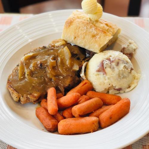 A plate featuring pork chop with gravy, mashed potatoes, glazed carrots, and a bread roll topped with butter.