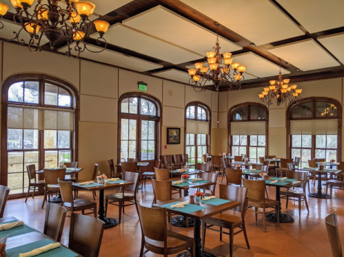 A spacious restaurant interior with wooden tables, chairs, and chandeliers, featuring large windows and a warm ambiance.