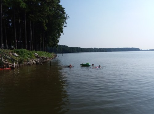 Two people swim in a calm lake surrounded by trees, with a green float nearby and a staircase leading to the water.