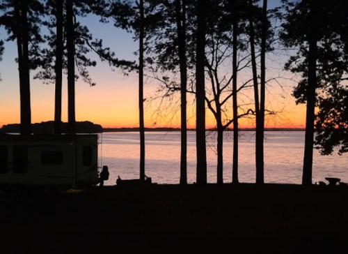 Silhouette of trees by a lake at sunset, with a camper in the foreground and vibrant colors in the sky.