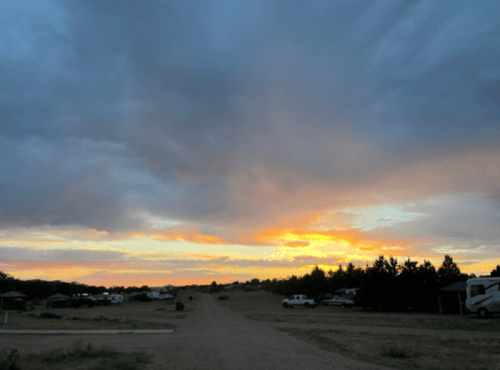A scenic sunset over a dirt road, with clouds and a hint of orange and yellow in the sky, surrounded by trees and RVs.
