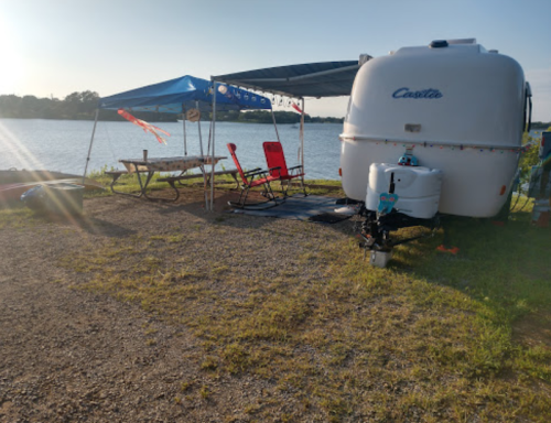 A cozy camper by a lake, with a picnic table, chairs, and a blue canopy set up for relaxation.