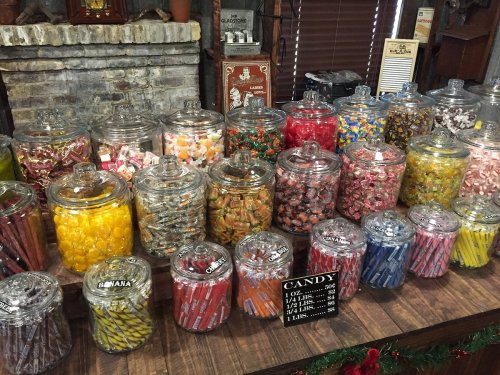 A colorful display of various candies in glass jars on a wooden table, with a rustic brick backdrop.
