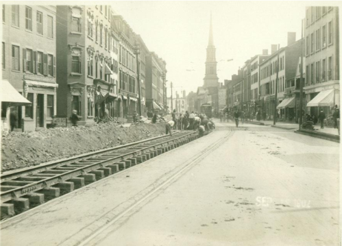 Historic street scene with workers laying tracks, buildings lining the street, and a church steeple in the background.