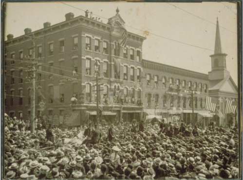 A large crowd gathers in front of historic buildings, with flags displayed and a church steeple in the background.