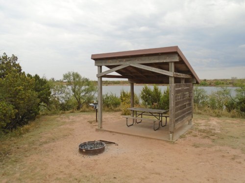 A wooden picnic shelter with a table near a fire pit, overlooking a calm lake and surrounded by greenery.