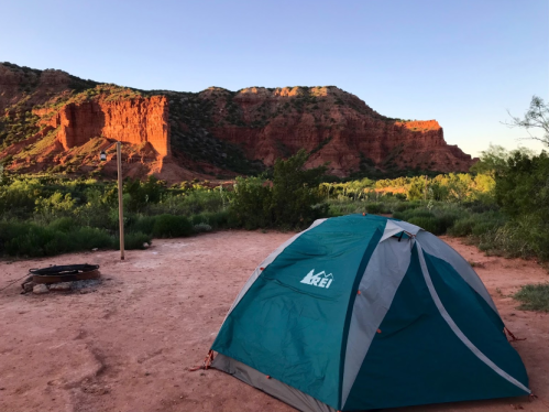 A teal tent set up in a sandy area with red rock formations and greenery in the background during sunset.