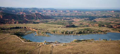 A panoramic view of a serene lake surrounded by rolling hills and red rock formations under a clear sky.