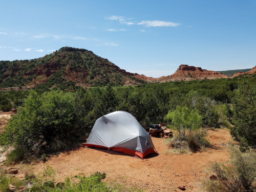 A tent set up in a desert landscape with green shrubs and rocky hills under a clear blue sky.