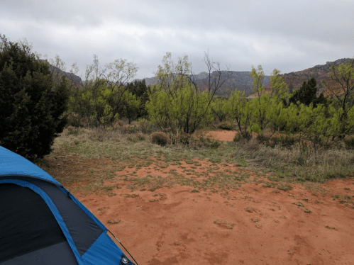 A blue tent in a grassy area surrounded by green shrubs and mountains under a cloudy sky.
