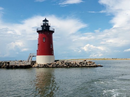 A red and white lighthouse stands on a rocky shore under a blue sky with scattered clouds, reflecting in the water.
