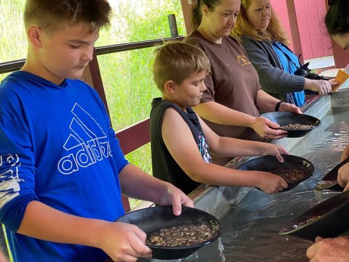 Children and adults panning for gold at a table outdoors, focused on their activity.