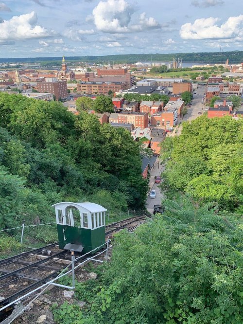 A funicular railway descends a hillside, overlooking a cityscape with greenery and clouds in the background.
