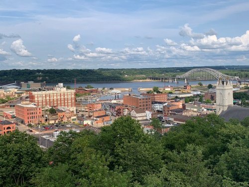 A scenic view of a riverside town with buildings, greenery, and a bridge under a partly cloudy sky.