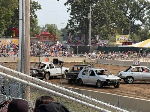 A demolition derby scene with damaged cars on a dirt track, surrounded by a large crowd at a fair.