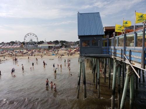 A beach scene with people swimming and sunbathing, featuring a pier and a ferris wheel in the background.