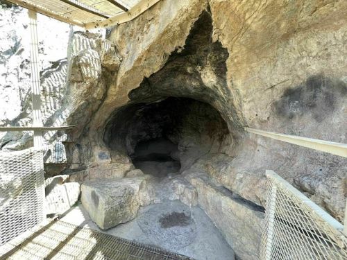 A rocky cave entrance with a metal railing, surrounded by natural stone formations and a dimly lit interior.