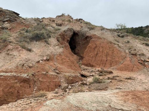 A rocky hillside with reddish soil and a cave-like opening, surrounded by sparse vegetation under a cloudy sky.