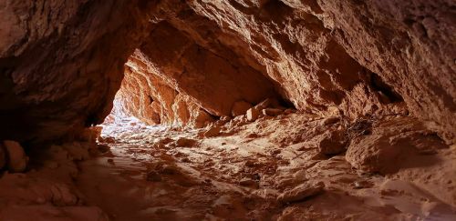 A rocky cave entrance with light streaming in, illuminating the sandy floor and rugged walls.