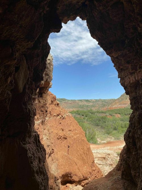 View from a cave opening, showcasing a blue sky and rugged landscape with red rock formations and greenery.