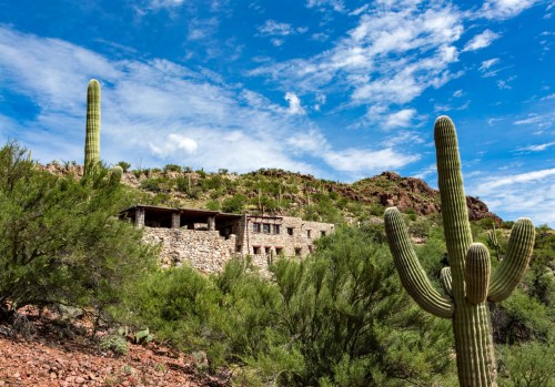 Stone building nestled in a desert landscape with tall cacti and a blue sky dotted with clouds.