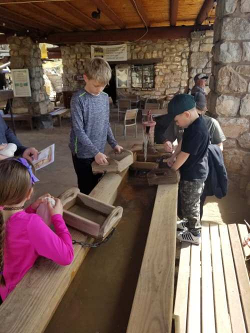 Children engaged in a hands-on activity at a wooden table, working with tools and materials in a rustic setting.
