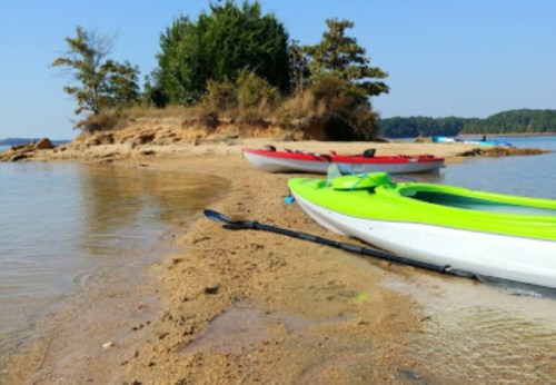 Two kayaks, one green and one red, are beached on a sandy shore with trees in the background and calm water.