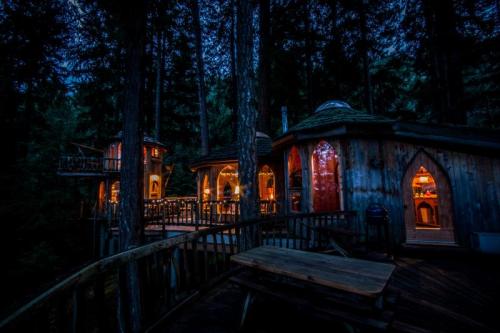 A cozy treehouse illuminated at night, surrounded by tall trees and a wooden deck. Warm lights glow from the windows.