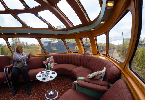 A woman sits in a cozy train car with large windows, as a train passes by outside.