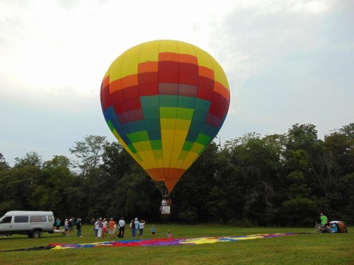 A colorful hot air balloon prepares for takeoff, surrounded by a crowd and a grassy field under a cloudy sky.