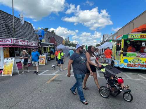 A busy street fair with food trucks, vendors, and people walking, including a couple with a stroller and a dog.