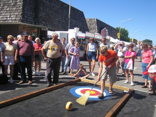 A crowd watches as a participant plays a game of curling on a street during a festival. Bright sunny day.